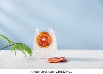 Cold Water With Red Orange And Ice In Glass On Pastel Blue Background. Refreshing Lemonade, Summer Drink, Cocktail, Soda. Empty Copy Space