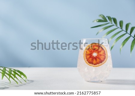 Cold water with red orange and ice in glass on pastel blue background. Refreshing lemonade, summer drink, cocktail, soda. Empty copy space