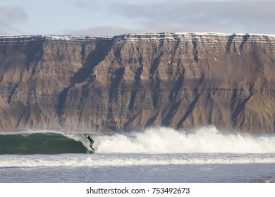 Cold water surfing in Iceland. Surfer catching a wave in the North Atlantic ocean. Snow covered mountain tops in the background.