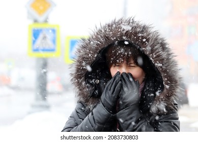 Cold weather and blizzard in winter city. Woman in fur hood standing on a street during snowfall and covering her face by hands in leather gloves
