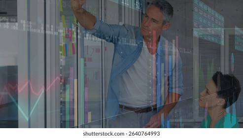 Collaborating colleagues marking sticky notes on glass partition in office, with marker and charts. Office, teamwork, innovation, digital, analytics, strategy, modern