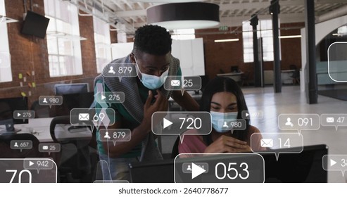 Collaborating masked colleagues analyzing social media stats on laptop in shared office, data icons. Collaboration, teamwork, modern office, digital analytics, professional environment, creative