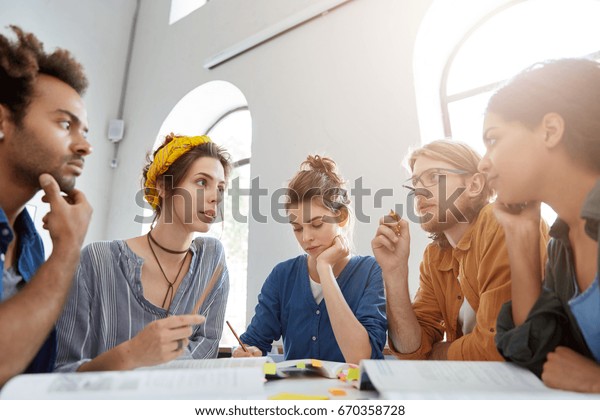 Collaboration and brainstroming concept. Portrait\
of interracial friends gathering together sitting at table in\
classroom surrounded with books having debates expressing their\
opinions and\
views.