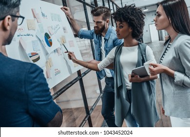 Collaboration is a key to best results. Group of young modern people in smart casual wear planning business strategy while young woman pointing at infographic displayed on the glass wall in the office