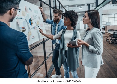 Collaboration is a key to best results. Group of young modern people in smart casual wear planning business strategy while young woman pointing at infographic displayed on the glass wall in the office