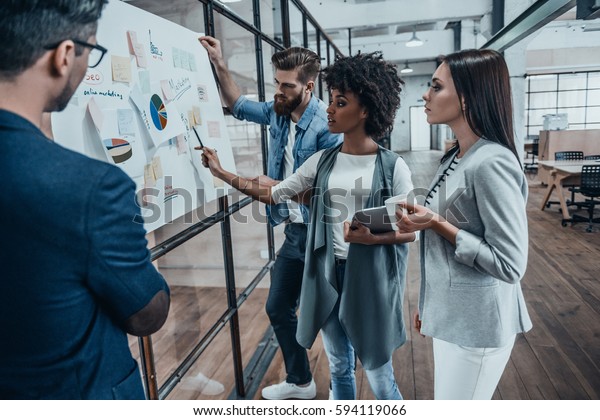 Collaboration is a key to best results. Group of\
young modern people in smart casual wear planning business strategy\
while young woman pointing at infographic displayed on the glass\
wall in the\
office