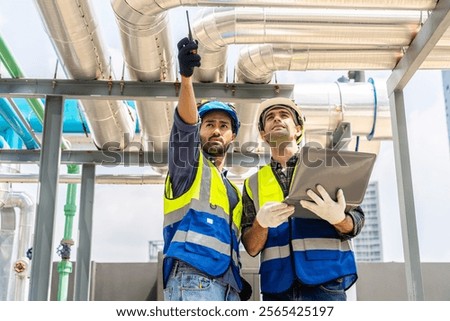 Collaboration, Two young construction workers discuss plans and use laptop computer while reviewing blueprints on a rooftop. They wear safety gear and oversee intricate piping systems in factory