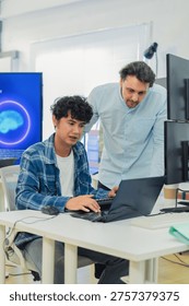 collaborative AI development team in modern tech office: young male developer coding on laptop while senior colleague leans over discussing code and data visualizations on multiple monitors with large AI Assistant display showing neural network graphics, symbolizing innovation machine learning programming teamwork coding collaboration.