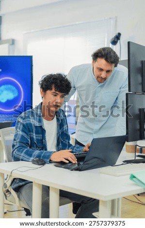 collaborative AI development team in modern tech office: young male developer coding on laptop while senior colleague leans over discussing code and data visualizations on multiple monitors with large AI Assistant display showing neural network graphics, symbolizing innovation machine learning programming teamwork coding collaboration.