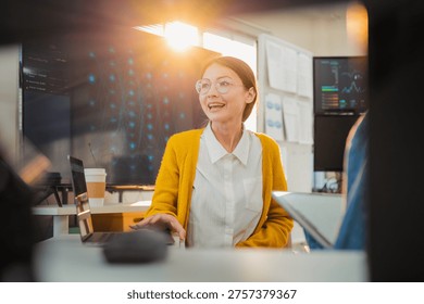 collaborative female AI developer in yellow sweater and glasses discusses code and neural network diagrams on large monitor while taking notes on tablet in modern tech office, with team members listening attentively, symbolizing innovation machine learning programming teamwork data analysis and agile development.