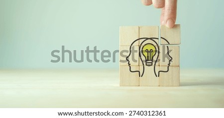 Collaborative Idea Generation. A hand stacks wooden blocks forming two people with a glowing lightbulb, symbolizing teamwork, brainstorming, innovation and shared problem solving.