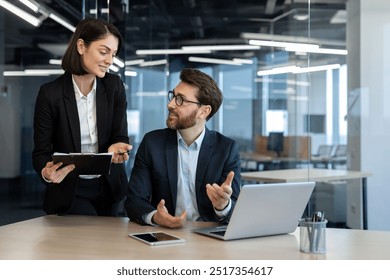 Colleagues engaged in discussion in modern office with laptop table. Woman holding documents, man seated. Interaction conveys teamwork, professionalism, corporate environment. - Powered by Shutterstock