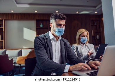 Colleagues With Face Masks Sitting In Business Space At An Official Business Meeting. Man Using A Laptop While A Woman In The Background Using A Tablet. Zoom Online Meeting, Coronavirus, COVID-19