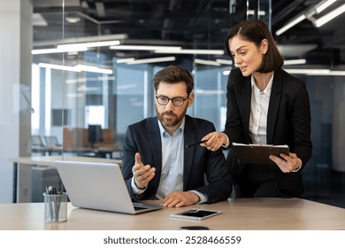 Colleagues in formal attire engaged in business meeting. Woman pointing at laptop screen, man contemplating solution. Office desk with laptop, smartphone, and documents enhancing collaborative - Powered by Shutterstock