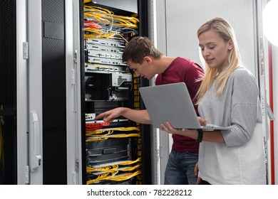 Colleagues Maintaining Server Room. Woman Holding Laptop While Man Is Checking The Cables