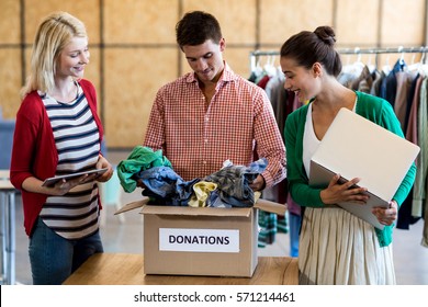 Colleagues using digital tablet and laptop while sorting clothes from donation box in the office - Powered by Shutterstock