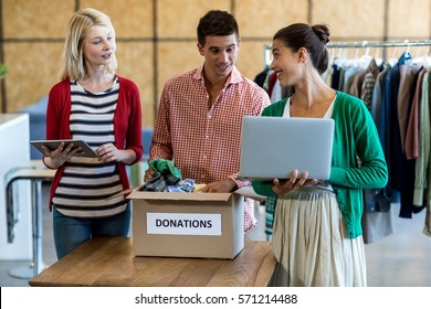 Colleagues using digital tablet and laptop while sorting clothes from donation box in the office - Powered by Shutterstock