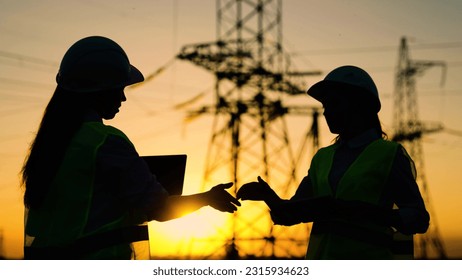 Colleagues women shake hands. Teamwork of power engineers in protective helmets, maintenance of power lines in outdoors. Two construction engineers work together on an electrical transmission line.
