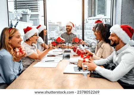 Colleagues at work give each other New Year's gifts while sitting at the table in the office. A group of people are sitting at a table in an office and exchanging gifts.                              