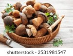 A collection of assorted fresh mushrooms is beautifully arranged in a woven basket, accompanied by a knife and vibrant herbs on a wooden table, showcasing nature