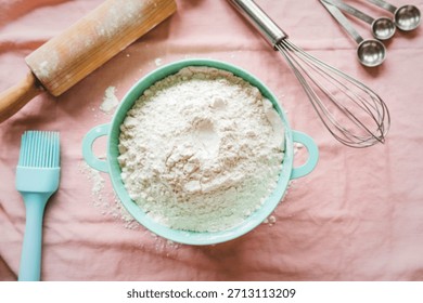 A collection of baking essentials including flour, a rolling pin, whisk, measuring spoons, and a brush, arranged on a pink cloth.