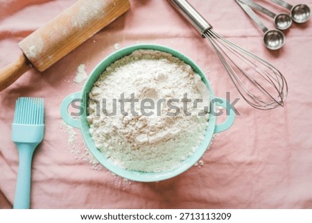 A collection of baking essentials including flour, a rolling pin, whisk, measuring spoons, and a brush, arranged on a pink cloth.