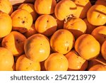 Collection of bright orange persimmons, displayed in a outdoor market under sunlight. The fruits are stacked closely together, showcasing their round and smooth texture