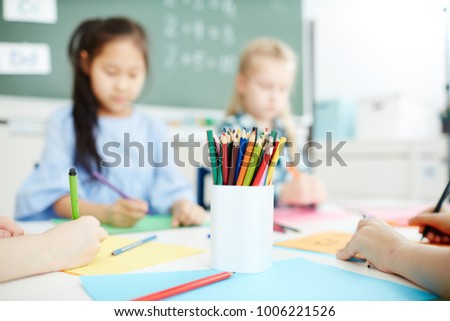 Collection of colorful crayons in pencilbox and group of kids drawing by table