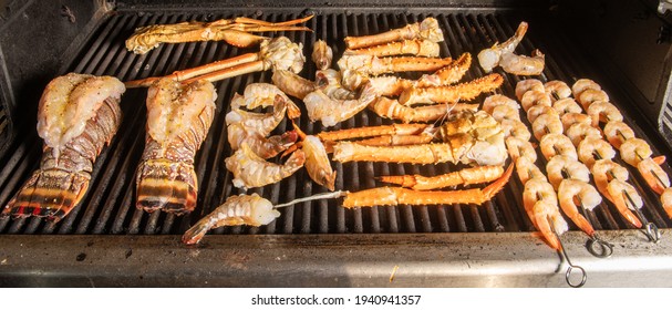 A Collection Of Different Kinds Of Seafood Bbq Cooking On A Grill With Lobster Tail That Is Saddleback, Tiger Shrimp, Shrimp On Skewers, Alaskan Snow Crab And Alaskan King Crab