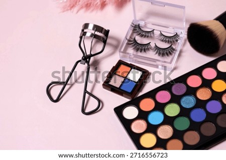A collection of eye makeup essentials including an eyeshadow palette, false lashes, eyelash curler, and a makeup brush on a light background.