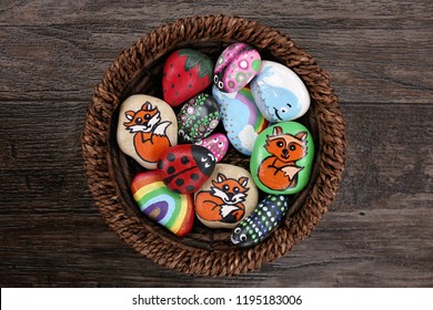 A collection of fun, handpainted, colorful cartoon rocks are together in a wicker basket, on a wooden plank background. - Powered by Shutterstock