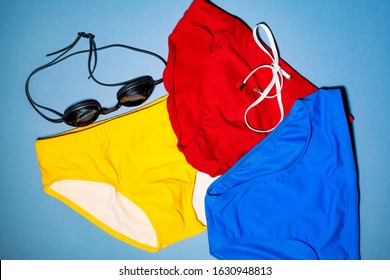 A Collection Men's Swim Briefs In Yellow, Red And Blue, And A Pair Of Swim Goggles, All On A Blue Background.