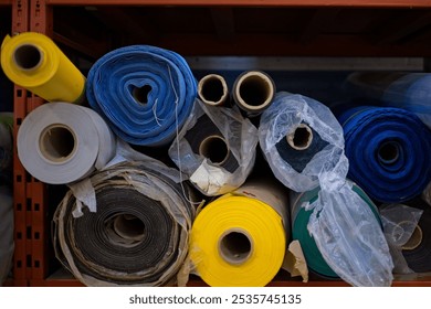 A collection of rolled fabric in various colors, including blue, yellow, green, and gray, neatly arranged on a shelf. The fabrics are wrapped in plastic and cardboard tubes with vibrant colors. - Powered by Shutterstock