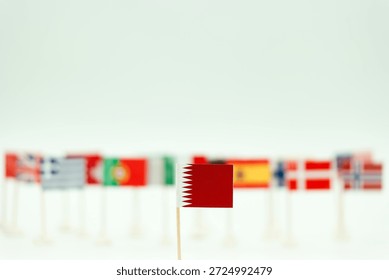 A collection of small flags from different countries is arranged on a flat surface. The red flag of Qatar is in the foreground while other flags form a blurred background.