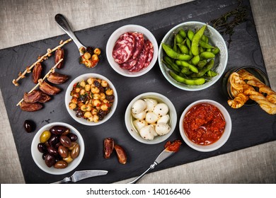 Tapas-Speisen wie z.B. Datteln, Kichererbensalat, Salami, frischer Mozzarella, Espamame und Oliven auf einem Schieferbrett.