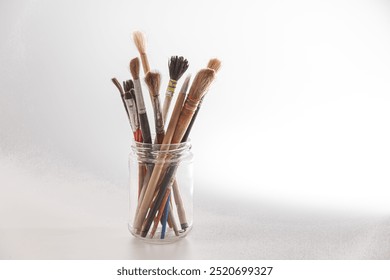 A collection of various paintbrushes stands upright in a clear glass jar, illuminated by natural light that enhances their textures and colors, showcasing the tools of an artist. - Powered by Shutterstock