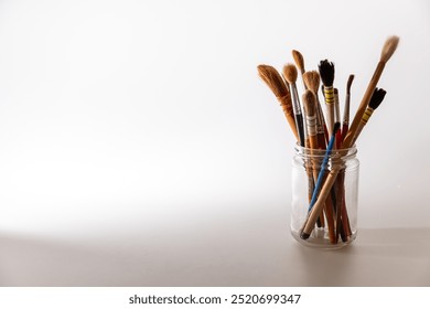 A collection of various paintbrushes stands upright in a clear glass jar, illuminated by natural light that enhances their textures and colors, showcasing the tools of an artist. - Powered by Shutterstock