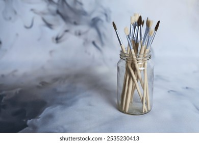 A collection of various paintbrushes with wooden handles displayed in a clear glass jar. The soft, abstract background creates a creative, artistic atmosphere. - Powered by Shutterstock