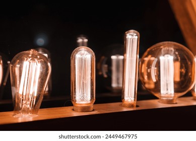 Collection of various shaped illuminated light bulbs emitting warm, amber glow on wooden surface. Filaments resemble  squirrel cage  type. Dark background creates cozy atmosphere. Location  Unknown. - Powered by Shutterstock