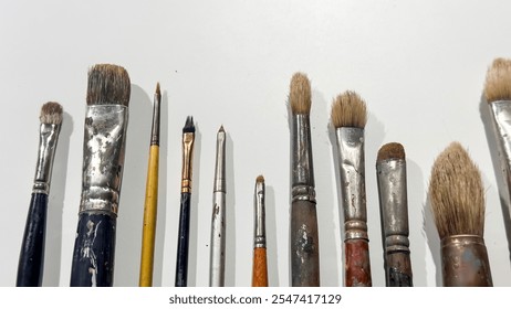 A collection of well-used paintbrushes of various sizes and shapes arranged horizontally on a plain white background. The bristles show signs of wear, reflecting creative use in painting projects. - Powered by Shutterstock