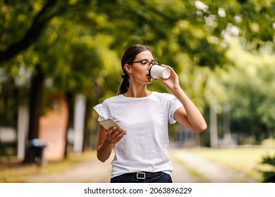 A College Girl Drinking Takeaway Coffee And Using Her Phone While Standing In The Campus On A Break Between Lectures. A College Girl In Campus