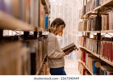 College girl reading and having toughts about the book while standing in the library between the bookshelves - Shutterstock ID 2267340449