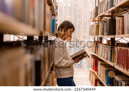 College girl reading and having toughts about the book while standing in the library between the bookshelves