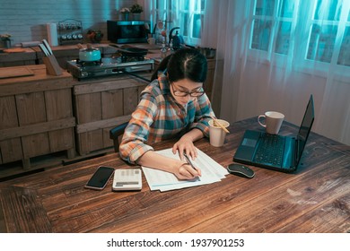 College Girl Student Stay Up Late Sitting At Table In Dark Home Kitchen At Night. Young Asian Korean Woman Enjoy Cup Of Instant Noodles Doing Homework And Preparing For Exam With Laptop Computer.