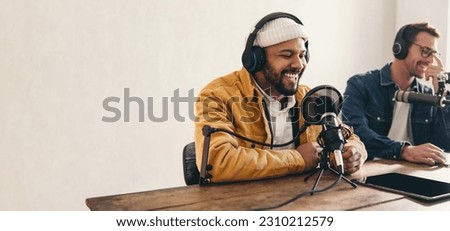 College radio dj smiling while speaking into a microphone in a studio. Happy young man co-hosting an audio broadcast with a guest. Two young content creators recording an internet podcast.