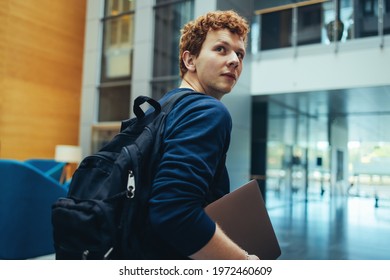 College student carrying his bag and laptop in campus. Young man turning back over his shoulder and walking in college campus. - Shutterstock ID 1972460609