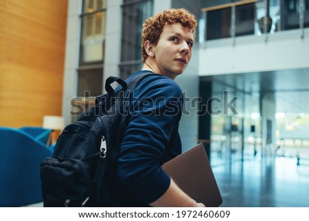 College student carrying his bag and laptop in campus. Young man turning back over his shoulder and walking in college campus.