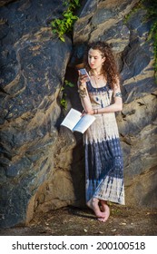 College Student. Dressing In Long Dress, Chunky Chain Bracelet, Arm Cuff Bracelet, Barefoot, A Teenage Girl With Curly Long Hair Is Standing Against Rocks, Holding A Book, Texting On Cell Phone.  