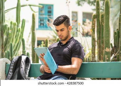 College Student Sitting On A Bench, Looking At His Tablet Computer