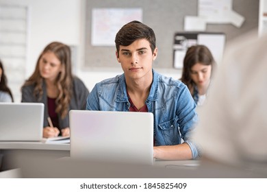 College Student Studying In Library With His Classmates In Background. High School Young Man Using Laptop For School Work During Computer Lesson. Portrait Of Satisfied University Guy Looking At Camera